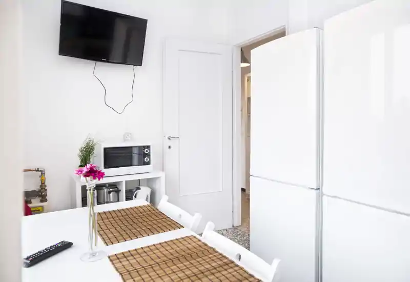 Bright compact kitchen with a white refrigerator, dining table with placemats and a mounted TV; neat and modern layout ideal for short stays.