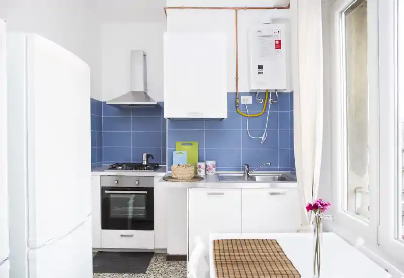 Bright compact kitchen with blue tile backsplash, gas hob and built-in oven, double sink, fridge and a small dining table by the window — clean modern styling ideal for a city apartment.
