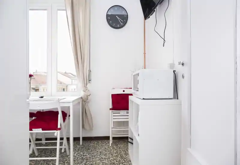 Compact kitchenette with a small dining table by a bright window; white furniture and red cushions create a neat, welcoming space suitable for short stays.