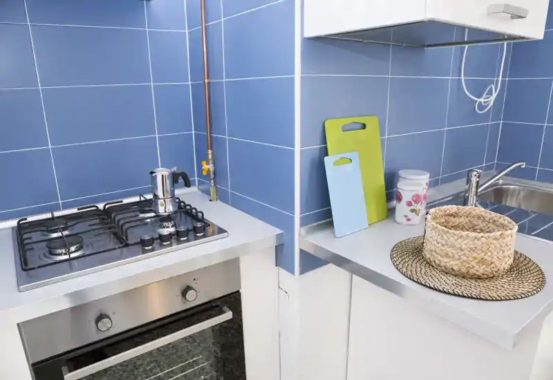 Compact modern kitchen with a gas hob, stainless-steel oven, sink and blue tiled backsplash. Small decorative details like cutting boards and a woven basket add warmth while keeping a clean, functional layout.