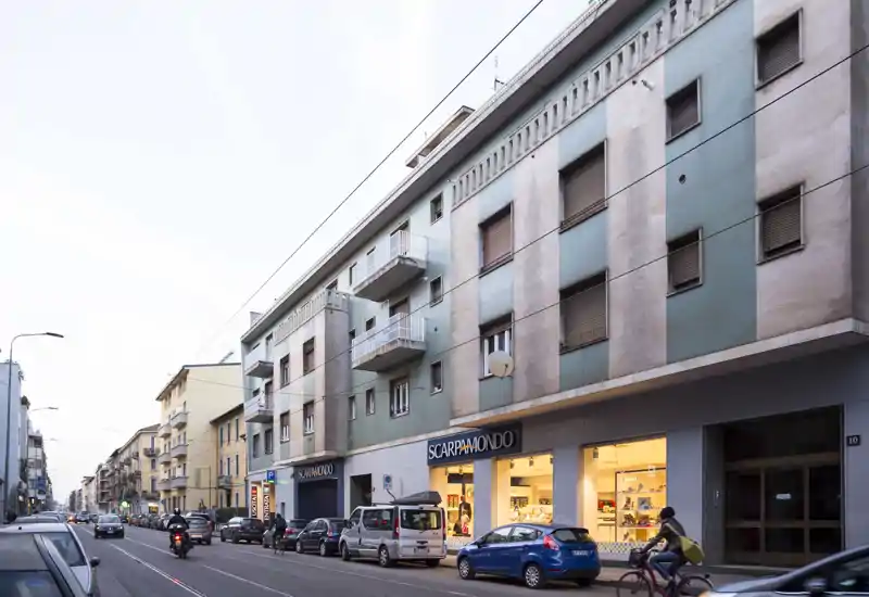 Street-level photo of a mixed-use building façade with ground-floor retail (ScarpaMondo), parked cars and a cyclist. Good evening light and clear architectural details.