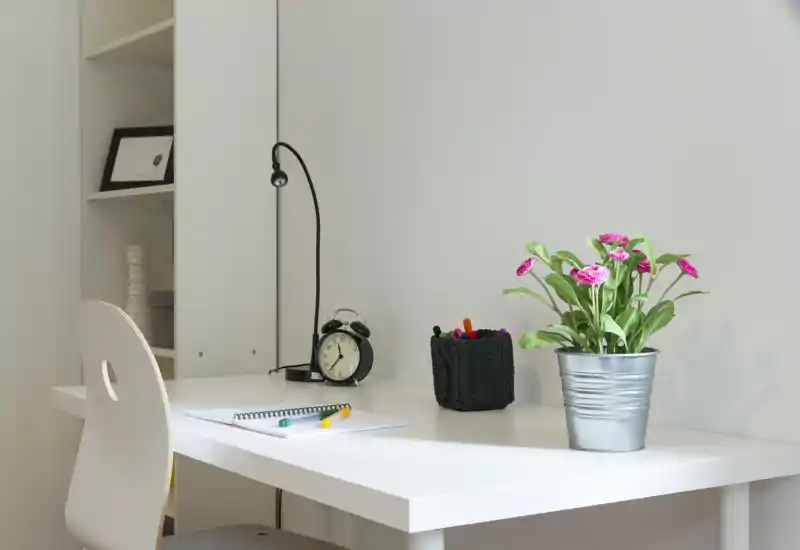 Angolo scrivania luminoso e minimal con tavolo bianco, sedia, lampada da ufficio, cancelleria e pianta in vaso — perfetto per lavoro o studio da remoto.