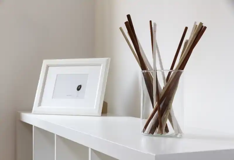 Un piccolo allestimento decorativo su una mensola bianca: una stampa incorniciata appoggiata al muro e un vaso di vetro trasparente con diversi bastoncini di legno. L'inquadratura mostra dettagli di styling, non un'intera stanza.
