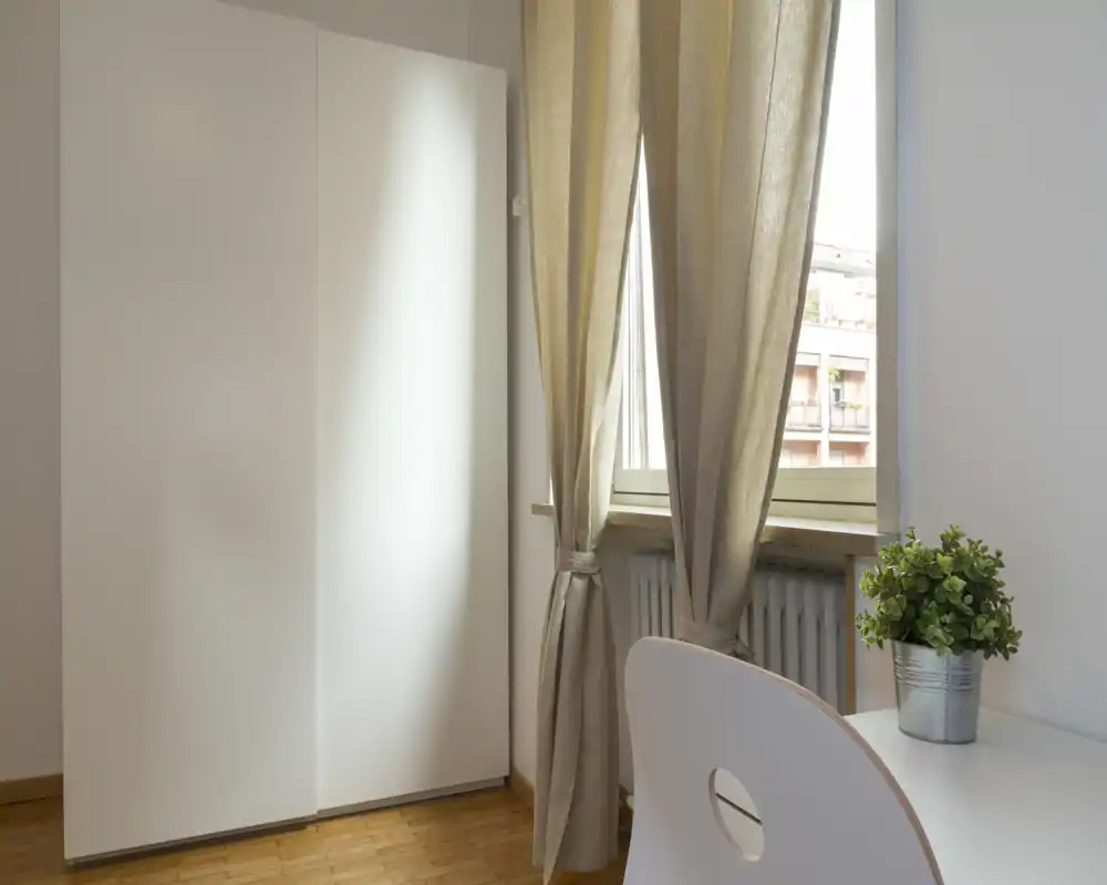 Angolo camera luminoso e minimal con un armadio bianco, scrivania e sedia vicino a una finestra con tende; stile semplice e pulito con una piccola pianta che crea un tocco accogliente.
