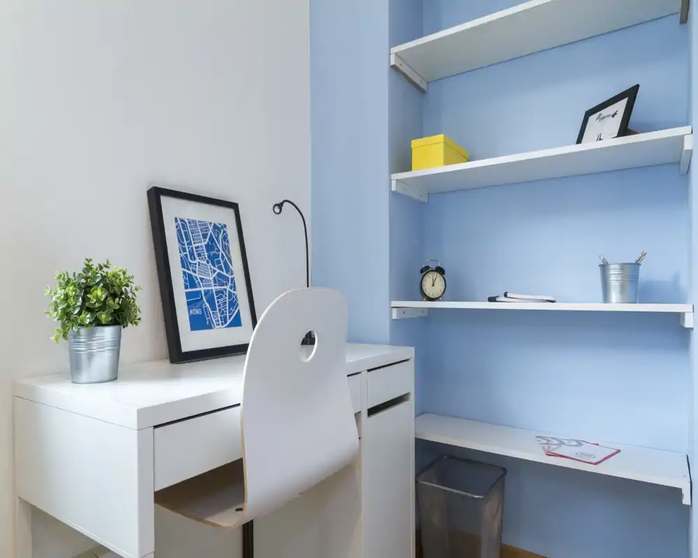 Angolo studio compatto e moderno con scrivania bianca, sedia e mensole integrate su una parete color azzurro; arredato con cura con una piccola pianta, una stampa incorniciata e accessori ordinati — ideale per smart working o studio.