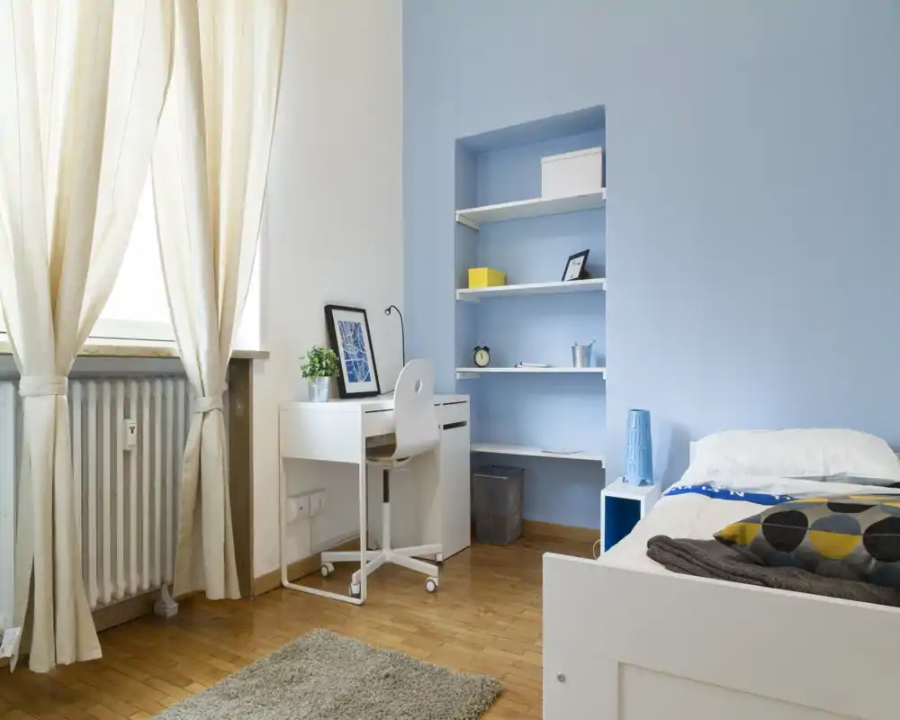 Camera da letto luminosa e moderna con letto singolo, scaffalatura a muro e una scrivania compatta vicino alla finestra — perfetta per uno studente o un inquilino singolo.