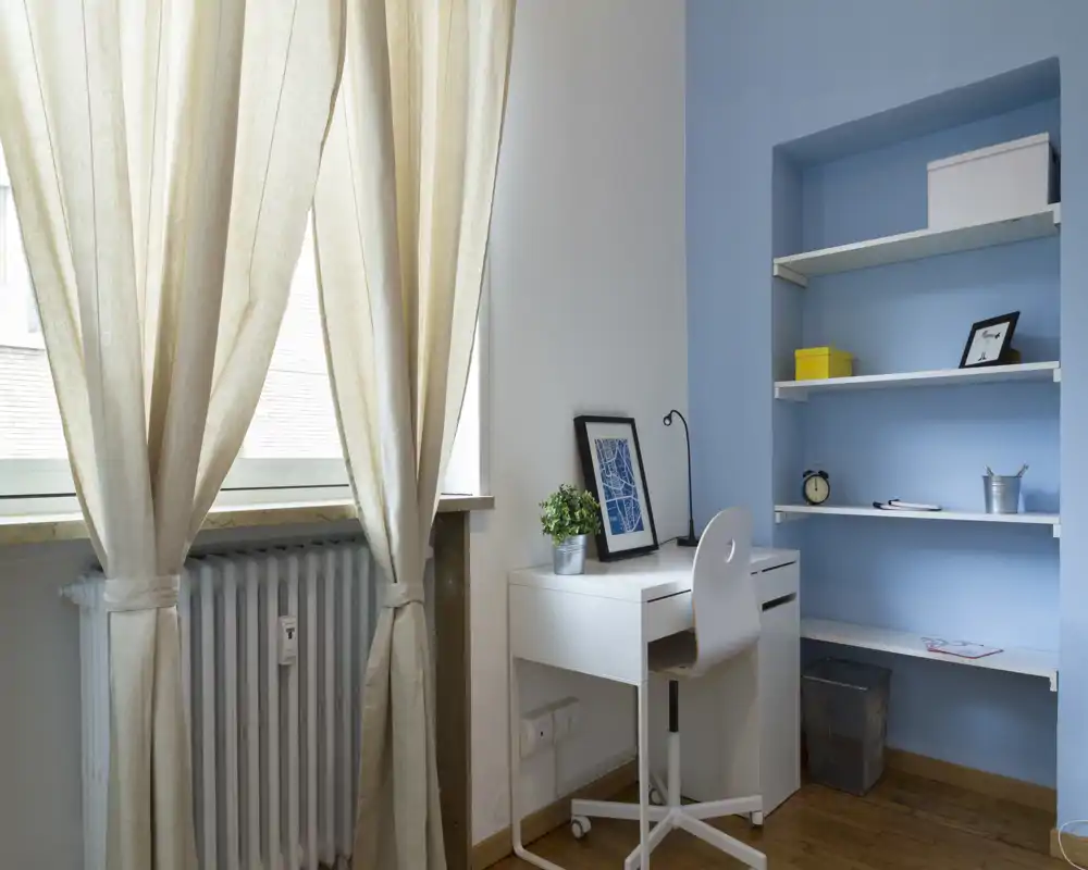 Angolo studio luminoso e ordinato con una piccola scrivania bianca, sedia ergonomica, scaffalatura blu incorporata e tende leggere che lasciano entrare la luce naturale — ideale per lavorare concentrati.