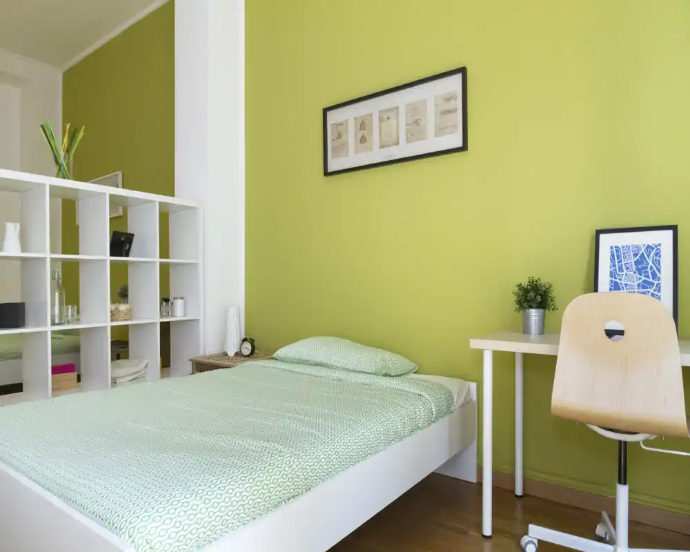 Camera da letto singola luminosa e moderna con letto ben rifatto, pavimento in legno chiaro, una piccola scrivania e scaffalatura decorativa — semplice e accogliente per un viaggiatore o uno studente.