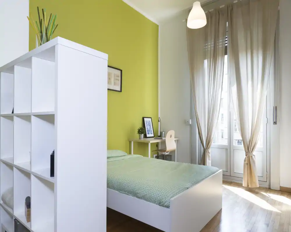 Camera da letto luminosa e moderna con letto matrimoniale, pavimento in legno chiaro e una piccola scrivania vicino alla finestra; tende leggere filtrano molta luce naturale e una libreria bianca funge da contenitore/divisorio.