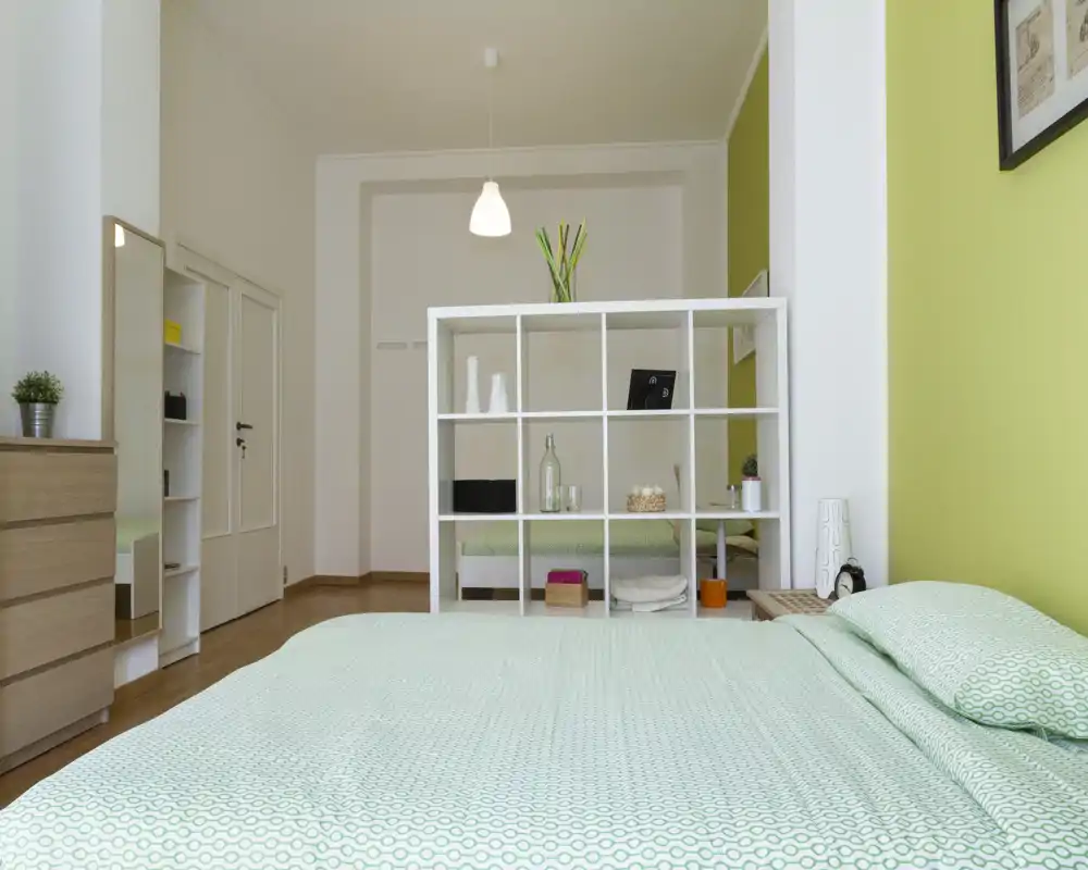 Camera da letto moderna e luminosa con letto in primo piano dai toni verdi, una libreria divisoria bianca e arredi minimal che creano un'atmosfera fresca e ariosa.