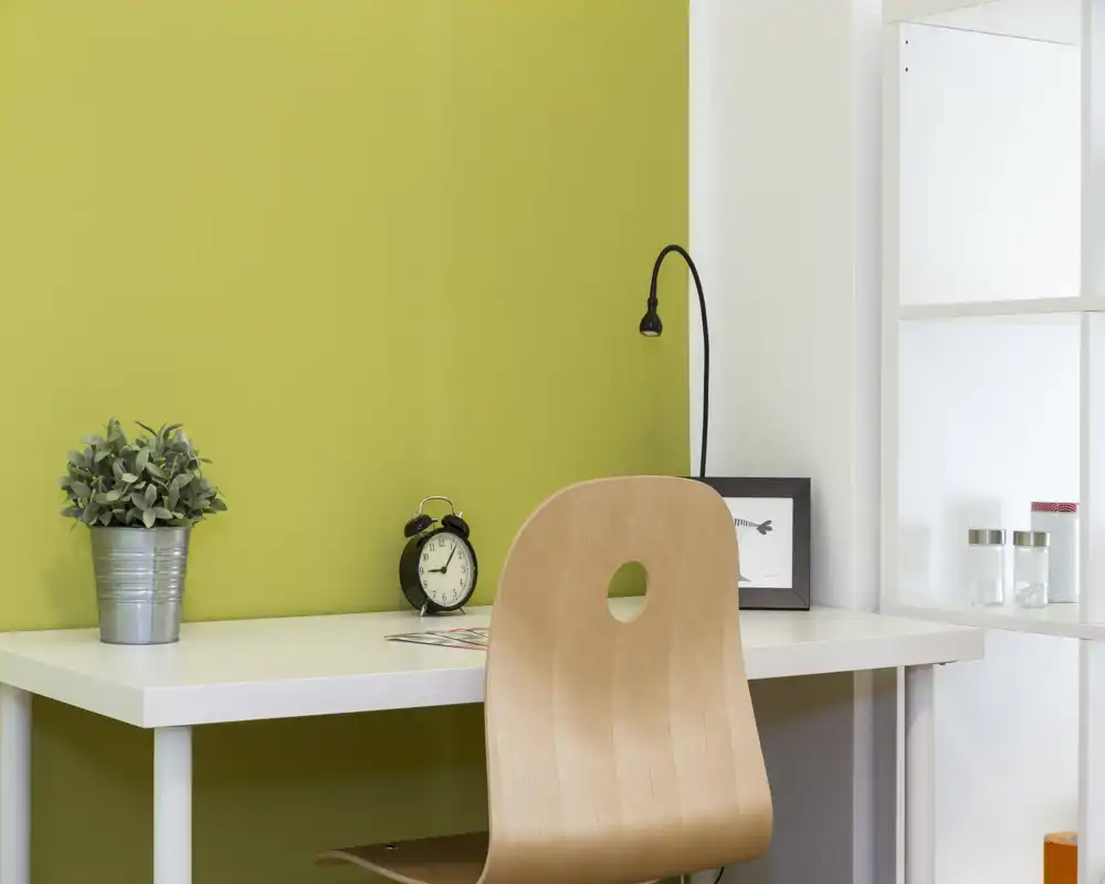 Una postazione ordinata con scrivania davanti a una parete verde, sedia in legno, piccola pianta, sveglia e lampada minimal: un angolo living/lavoro compatto e moderno.