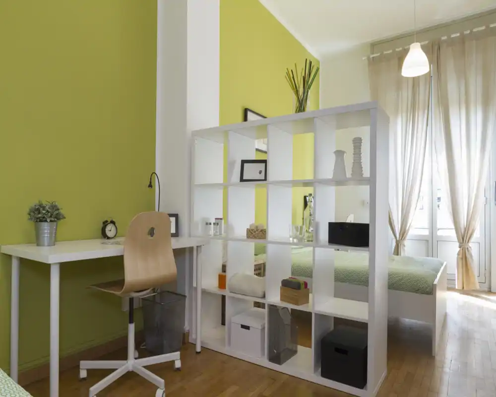 Camera da letto luminosa e ben arredata con una libreria a giorno bianca che separa il letto da un angolo studio; ideale per una persona o uno studente.