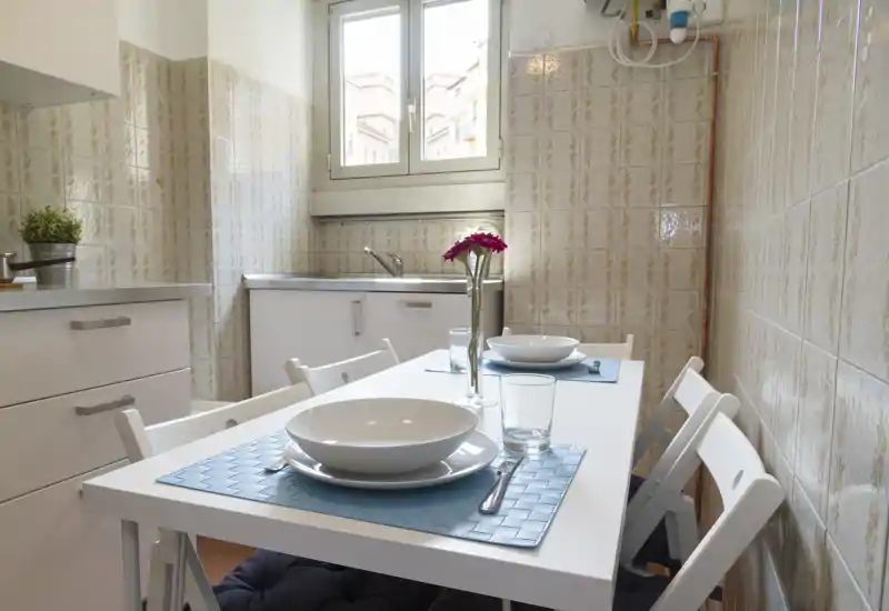 Cucina compatta e luminosa con un piccolo tavolo apparecchiato per due, luce naturale dalla finestra e mobili bianchi puliti — perfetta per un appartamento cittadino accogliente.