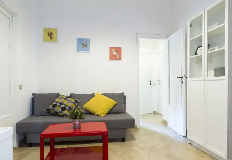 Area living luminosa e ordinata con divano grigio compatto, cuscini colorati e tavolino rosso; ideale per un piccolo appartamento o monolocale.
