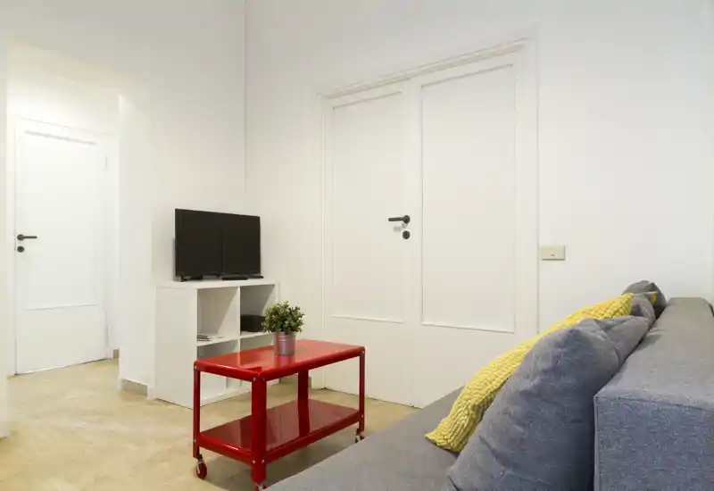 Soggiorno compatto e luminoso con divano grigio, cuscini gialli, un tavolino rosso su ruote e una piccola TV: stile pulito e moderno, ideale per affitti brevi.