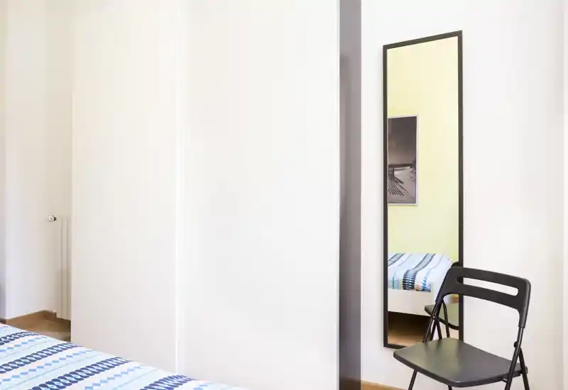 Camera da letto luminosa e minimale con biancheria a righe, un armadio e uno specchio a tutta altezza che riflette parte del letto; arredamento pulito e tranquillo, ideale per un annuncio.