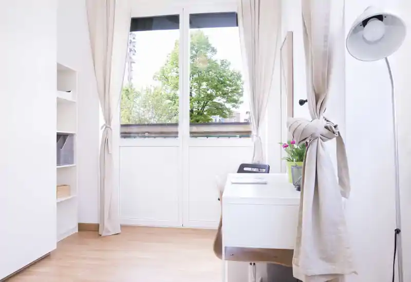 Zona giorno luminosa e minimal con ampia finestra che fa entrare luce naturale, una scrivania con sedia, scaffalatura semplice e tende leggere — perfetta come angolo lavoro in un appartamento compatto.