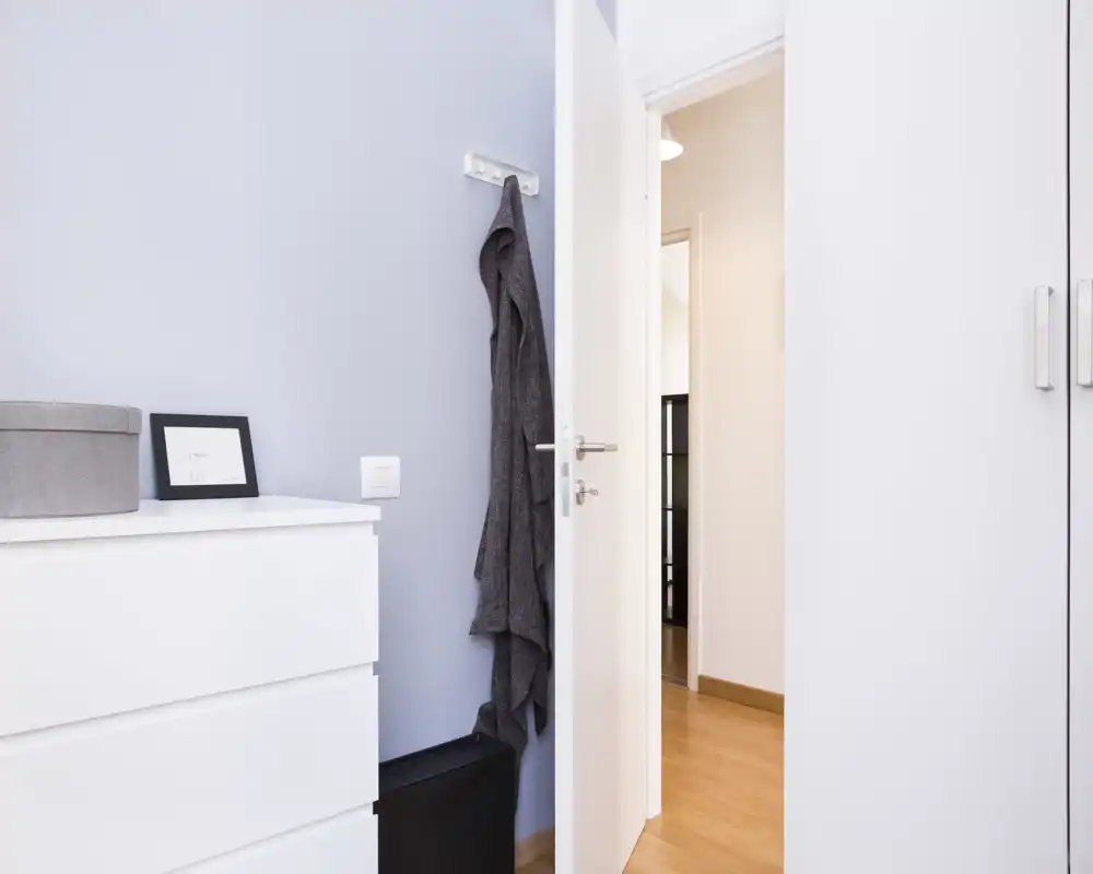 Entrada ordenada con perchero, cómoda blanca y una puerta entreabierta que conduce al apartamento. Luminosa, minimalista y bien organizada — ideal para mostrar el recibidor.