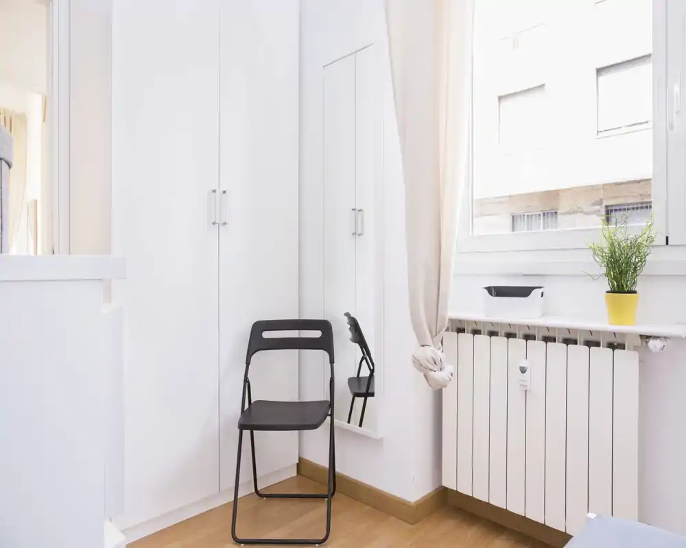 Pequeño rincón de estar luminoso con una silla plegable, un armario de altura completa y un alféizar con una planta — ambiente minimalista y limpio.