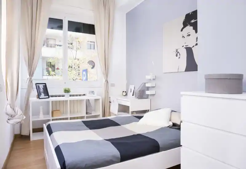 Dormitorio luminoso y bien decorado con cama doble arreglada, gran ventana con cortinas ligeras, escritorio y almacenamiento: ideal para una estancia urbana acogedora.