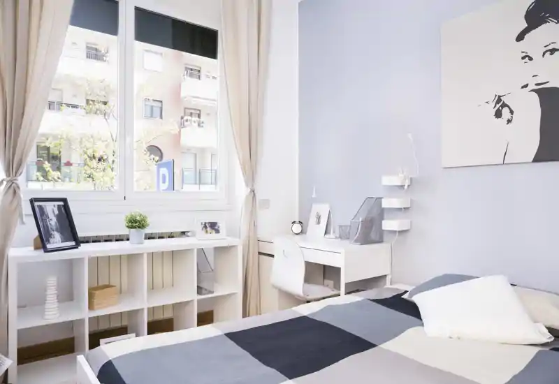 Dormitorio luminoso y bien amueblado con gran ventana, cama ordenada, escritorio y estantería — ideal para una persona o estudiante.