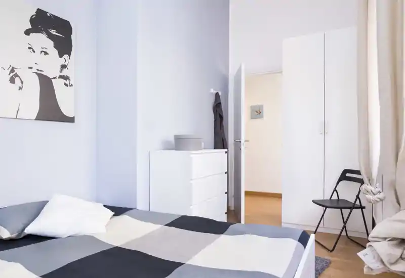 Dormitorio moderno y luminoso con una gran cama en primer plano, cómoda y armario blancos, tonos neutros y luz natural desde la ventana.