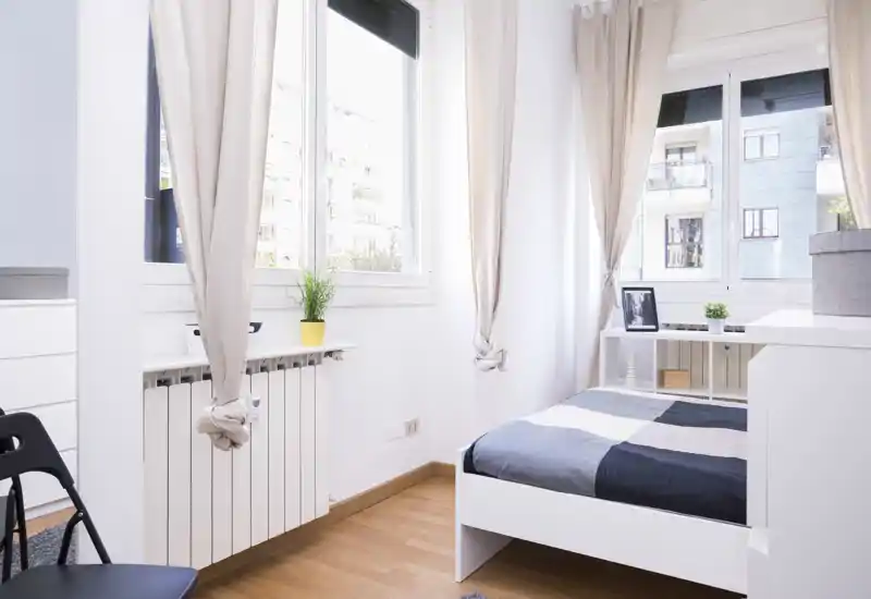 Dormitorio luminoso y minimalista con cama individual junto a grandes ventanas, suelo de madera clara y cortinas neutras que crean un ambiente moderno y luminoso.