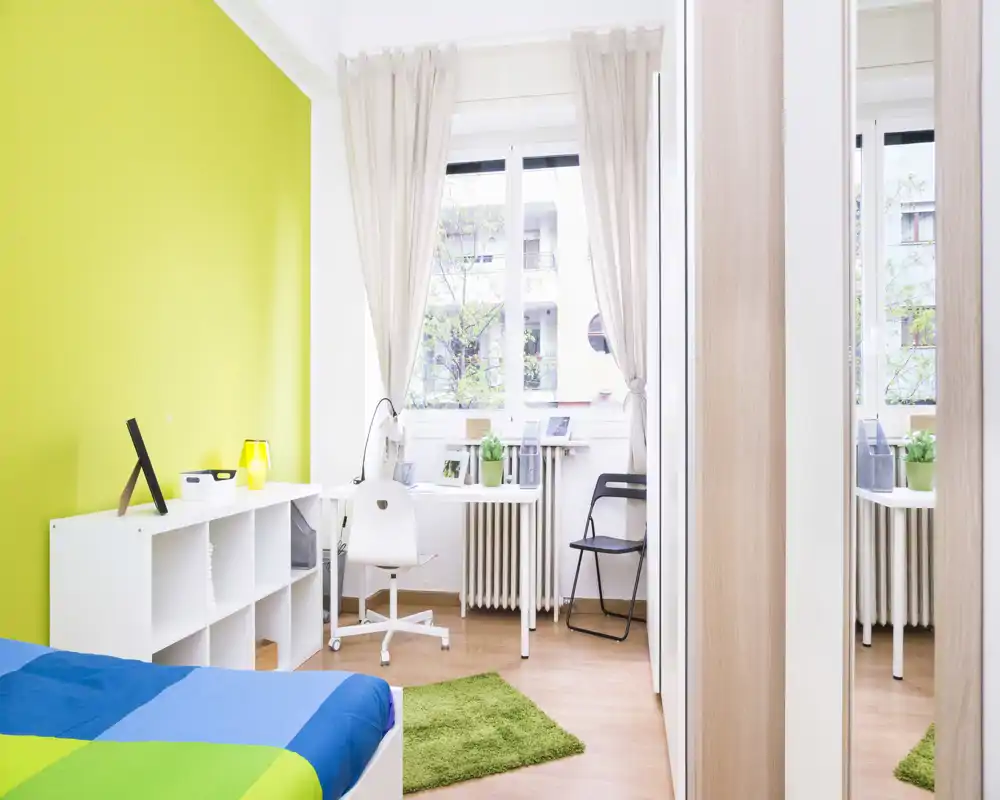 Camera da letto luminosa e moderna con letto colorato, scrivania vicino alla finestra e capiente libreria: perfetta per uno studente o single.