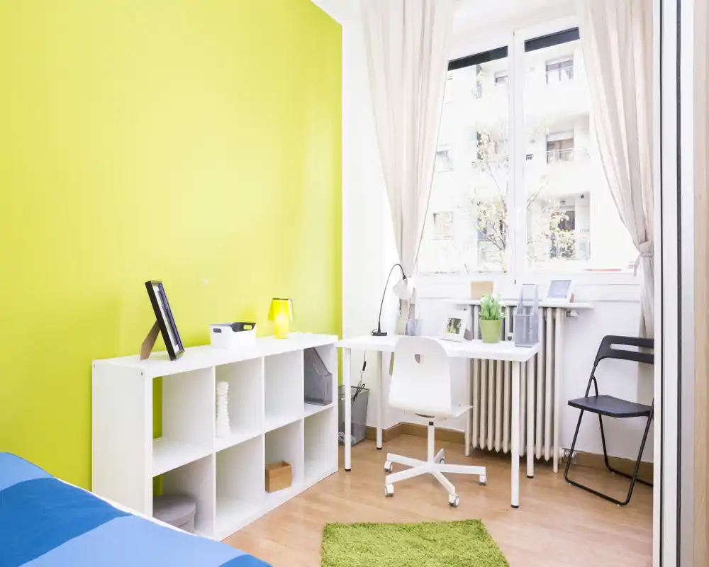 Camera luminosa e moderna con angolo studio: scrivania bianca vicino a una grande finestra, letto parzialmente visibile a sinistra, scaffalatura modulare e parete verde che crea un’atmosfera fresca e ariosa.