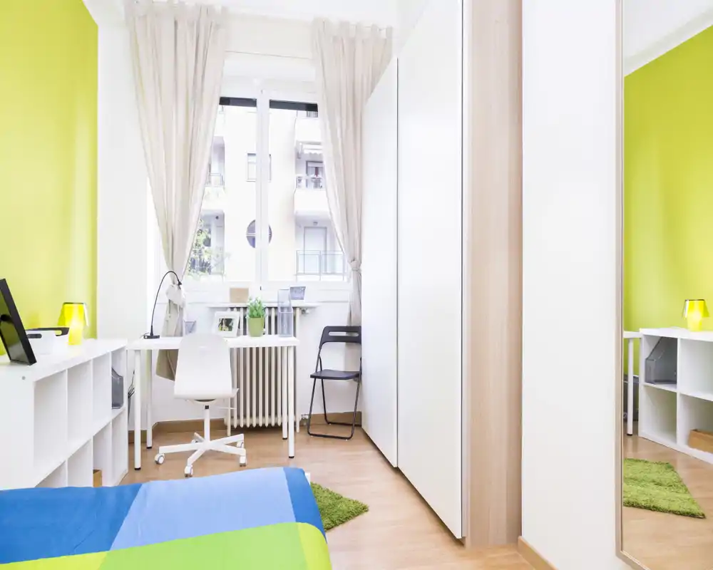 Camera da letto moderna e luminosa con letto colorato, scrivania vicino alla finestra e armadio a muro. La grande finestra illumina l'ambiente creando un'atmosfera fresca e accogliente.