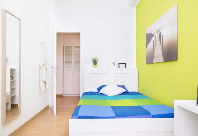 Camera da letto singola luminosa e moderna con telaio del letto bianco, biancheria blu-verde vivace e parete accentuata verde. Linee pulite, arredamento essenziale e uno specchio grande rendono la stanza ariosa e curata.