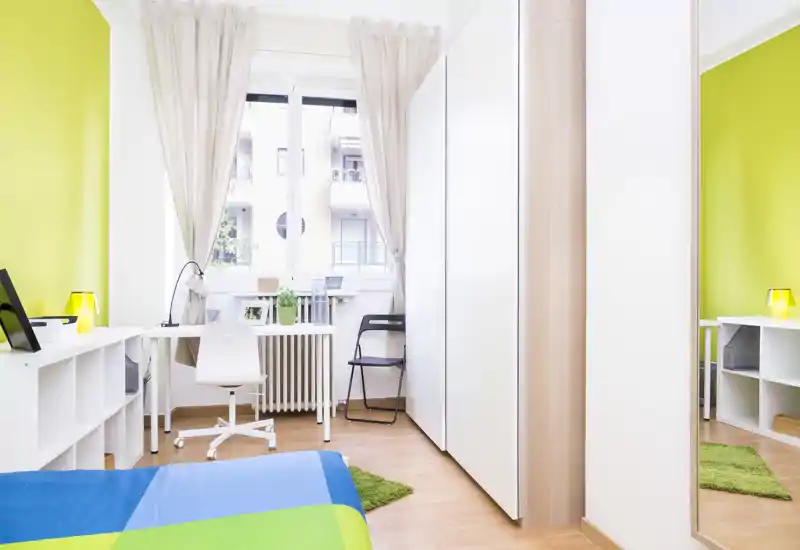 Camera da letto moderna e luminosa con letto colorato, scrivania vicino alla finestra e armadio a muro. La grande finestra illumina l'ambiente creando un'atmosfera fresca e accogliente.