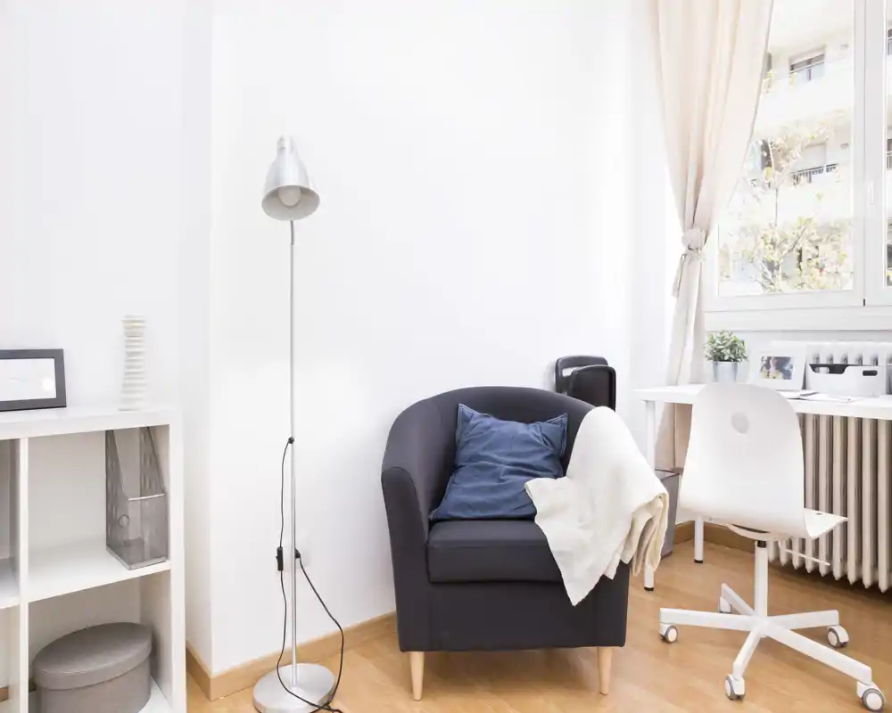 Angolo soggiorno luminoso e minimal con poltrona comoda, lampada da terra e una piccola scrivania vicino alla finestra — ideale per rilassarsi o lavorare leggermente.