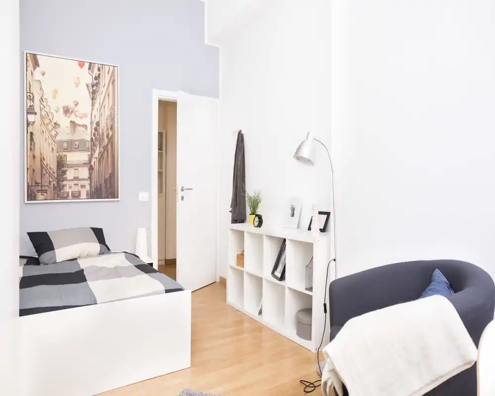 Camera da letto moderna e luminosa con un letto singolo ben rifatto, scaffalatura integrata e una comoda poltrona — perfetta per un soggiorno confortevole da solo.