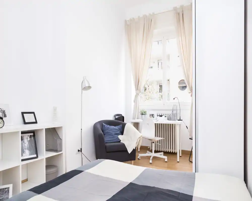 Camera da letto luminosa e ariosa con letto in primo piano, una poltrona accogliente e una scrivania vicino alla finestra — ideale per riposo e smart working.