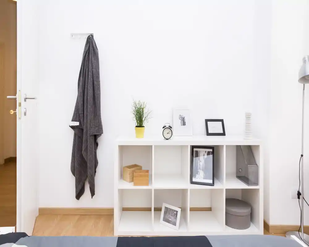 Angolo di camera da letto minimalista con un mobile bianco a cubi, cornici decorative e una pianta; parte del letto è visibile in primo piano, spazio luminoso e ordinato.