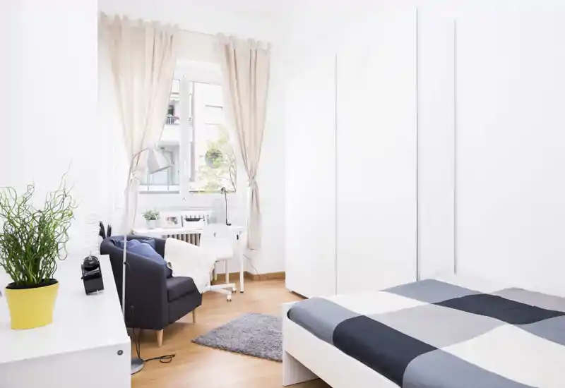 Camera da letto luminosa e moderna con un letto matrimoniale con piumone geometrico, un armadio bianco e un angolo lettura accogliente vicino alla finestra con scrivania e poltrona.