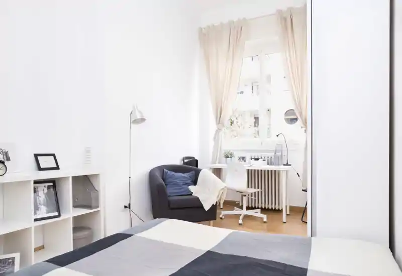 Camera da letto luminosa e minimale con letto ben rifatto in primo piano, una poltrona accogliente e una piccola scrivania vicino alla finestra che lascia entrare molta luce naturale.