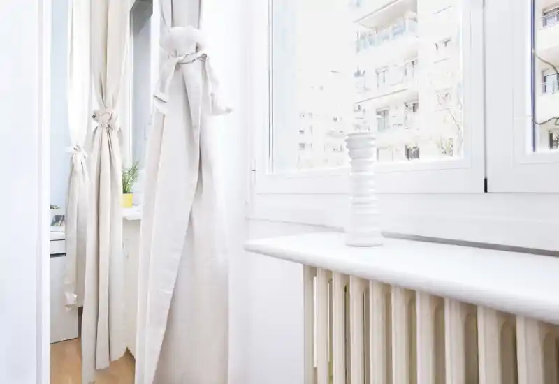 Area finestra luminosa con termosifone bianco e tende chiare; uno spazio minimale e pulito che affaccia su edifici vicini.