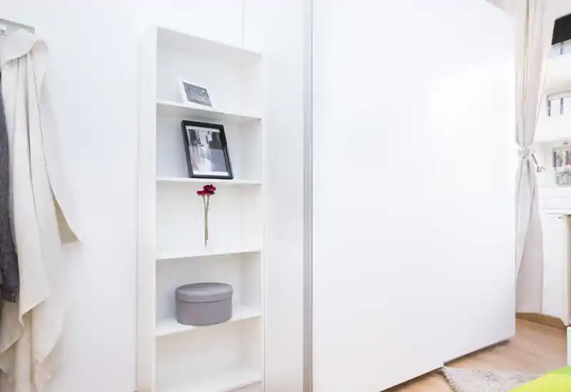 Angolo di camera minimal con una libreria alta bianca, porta scorrevole dell'armadio e una finestra che illumina l'ambiente; tessuti neutri e un piccolo tappeto creano un'atmosfera semplice e tranquilla.