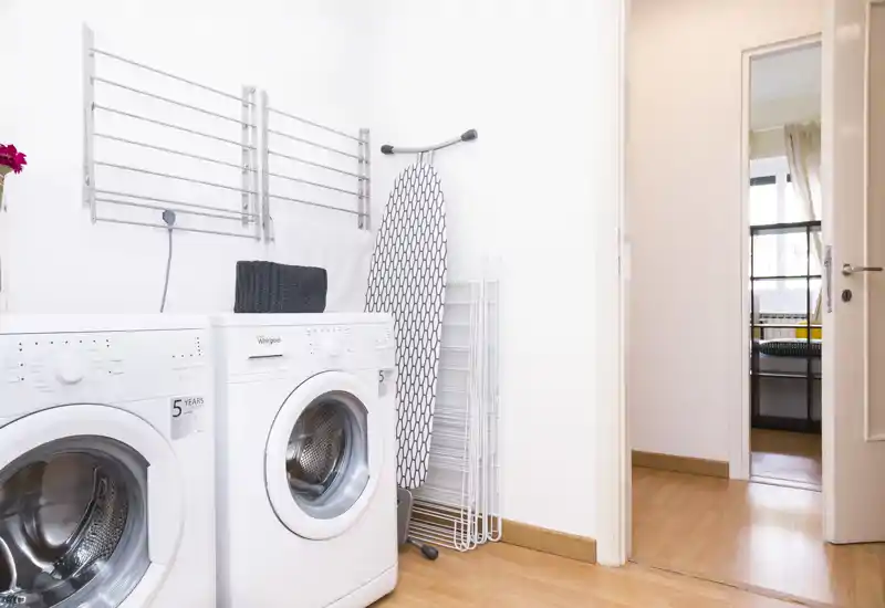 Area lavanderia luminosa e pulita con due lavatrici a carica frontale, asse da stiro e stendibiancheria — spazio funzionale perfetto per annunci di appartamenti.
