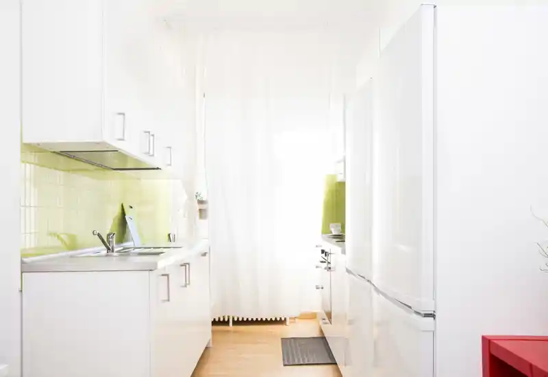 Cucina moderna e luminosa con mobili bianchi, paraschizzi in piastrelle verdi, lavello e frigorifero; spazio compatto, ben illuminato e ordinato, perfetto per annunci.
