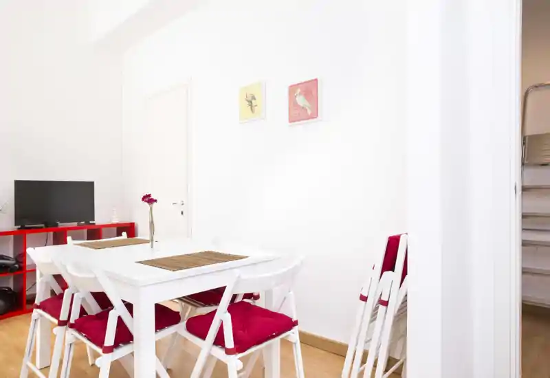 Zona pranzo luminosa e minimal con tavolo bianco, sedie pieghevoli bianche con cuscini rossi e un piccolo scaffale multimediale rosso — perfetta per un appartamento compatto.