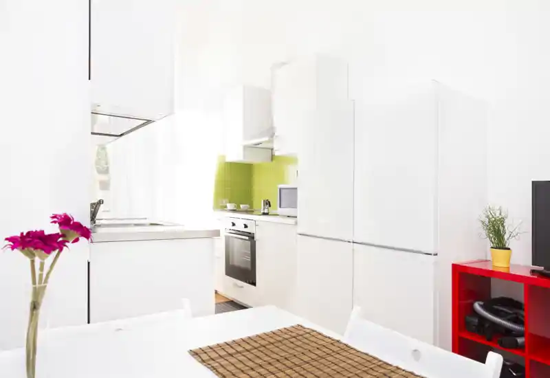 Cucina moderna e luminosa con mobili bianchi, forno incassato, grande frigorifero e paraschizzi in piastrelle verde lime; tavolo ordinato con fiori che dona un tocco fresco e accogliente.
