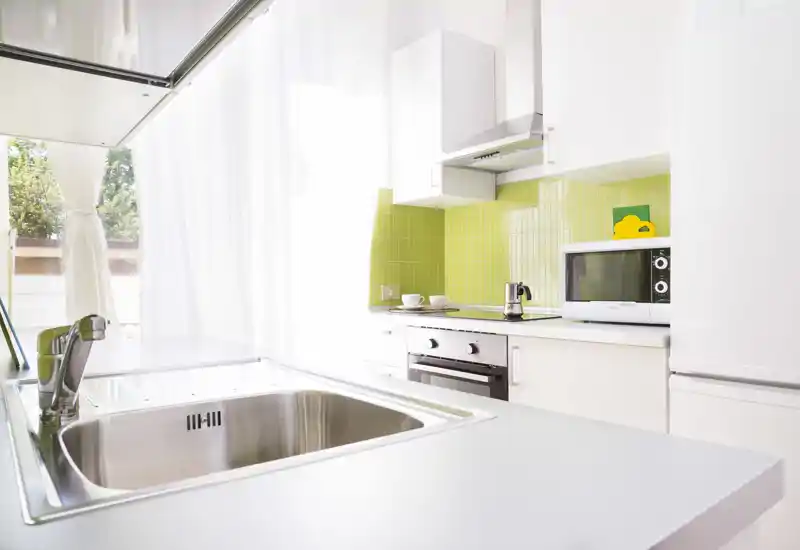 Cucina moderna e luminosa con lavello in acciaio inox, forno incassato e microonde, e un vivace paraschizzi in piastrelle verdi; ampia finestra con tende leggere che illumina lo spazio.