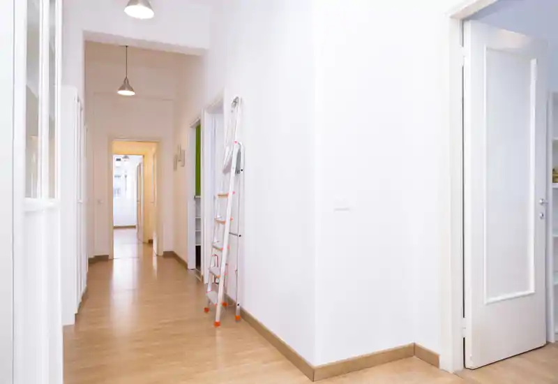 Corridoio luminoso e pulito con pavimento in legno e pareti bianche, dotato di lampade a sospensione e di una scala pieghevole appoggiata al muro.