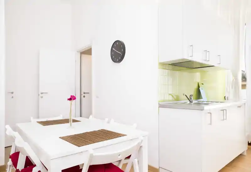 Zona pranzo luminosa e moderna con tavolo bianco per quattro accanto a una cucina compatta, caratterizzata da decorazioni semplici e luce naturale.