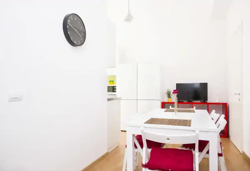Zona pranzo luminosa e compatta con tavolo bianco, sedie con cuscini rossi e piccolo mobile TV; arredo minimalista e luce naturale rendono l'ambiente fresco e accogliente.