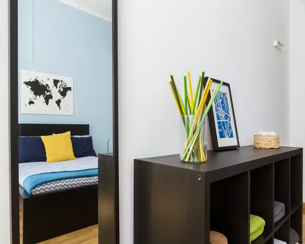 Camera da letto moderna visibile in gran parte attraverso uno specchio alto, con copriletto a tema, cuscini blu e un cuscino giallo; davanti un mobile a cubi con oggetti decorativi crea un gradevole angolo d'ingresso.
