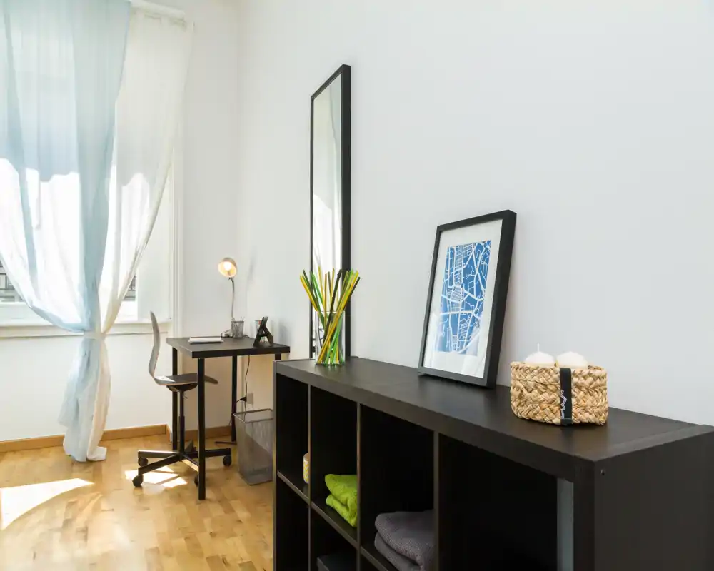 Zona giorno luminosa e minimal con una scrivania compatta, mobile a cubi e luce naturale da un’ampia finestra — ideale anche come angolo studio.